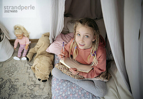 Lächelnd mit Buch sitzend neben Spielzeug im Zelt zu Hause