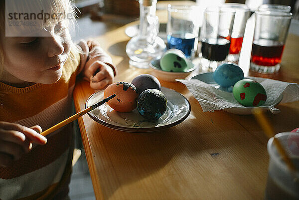 Girl decorating Easter egg with paintbrush at home