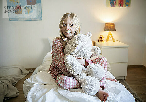 Smiling girl in pajamas sitting with teddy bear on bed at home