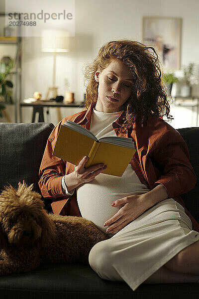 Expectant woman reading book sitting with dog on sofa at home