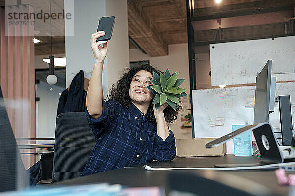 Lächelnde Geschäftsfrau  die im Büro ein Selfie mit einer Pflanze in Gesichtsnähe macht