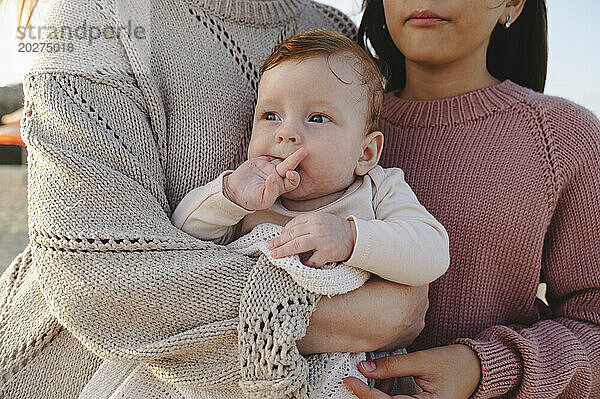 Niedliches kleines Mädchen in den Armen der Mutter in der Nähe ihrer Schwester am Strand