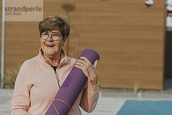Happy senior woman with exercise mat in front of brown wall