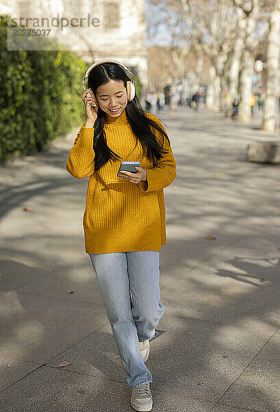 Lächelnde junge Frau trägt kabellose Kopfhörer und benutzt ein Smartphone