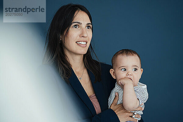 Thoughtful businesswoman carrying daughter in front of blue background