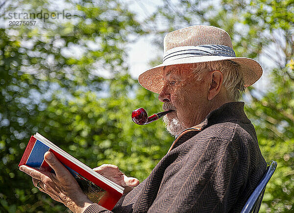 Älterer Mann raucht Pfeife und liest Buch im Garten