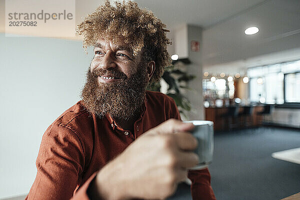 Lächelnder reifer Geschäftsmann mit Afro-Frisur hält Kaffeetasse in der Bürokantine