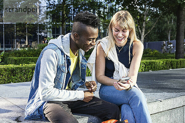 Lächelnde Frau teilt Smartphone mit Freund  der auf Bank sitzt