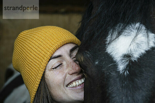 Glückliche Frau mit geschlossenen Augen neben geschecktem Pferd im Stall