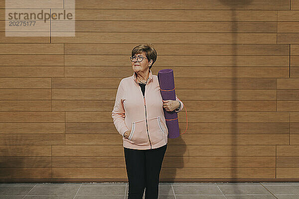 Ältere Frau mit Gymnastikmatte steht vor brauner Holzwand