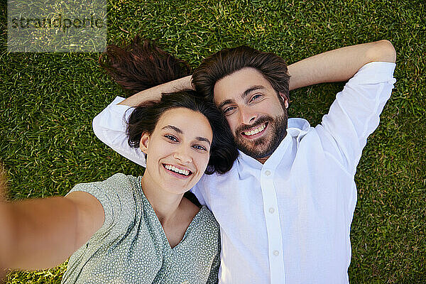 Lächelnde junge Frau macht ein Selfie mit einem Mann  der im Gras liegt