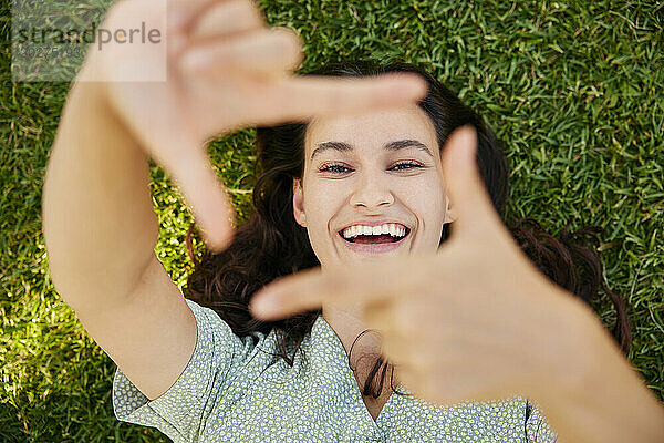 Happy woman looking through finger frame and lying on grass