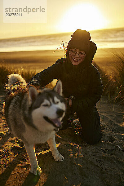 Smiling woman kneeling and petting Husky dog at beach