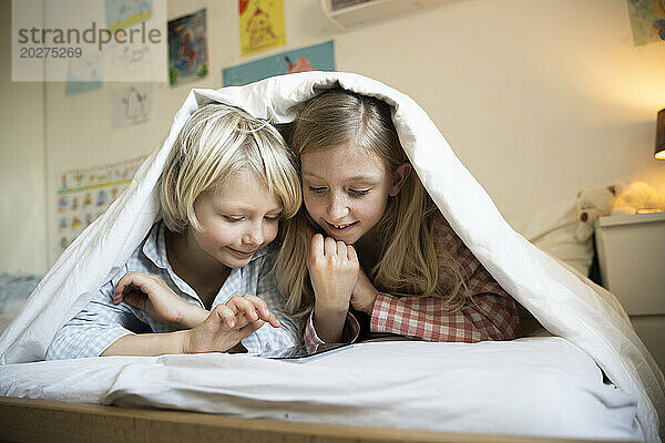 Bruder und Schwester schauen zu Hause auf das Smartphone und liegen unter der Decke im Bett