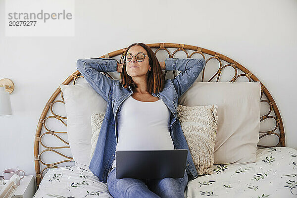 Pregnant freelancer resting on bed with hands behind head at home office