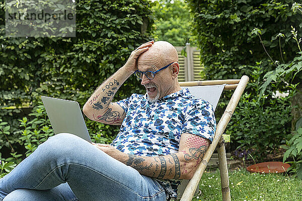 Überraschter Mann mit Kopf in der Hand blickt im Garten auf Laptop