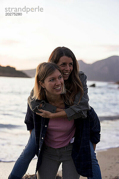 Carefree woman piggybacking friend on shore at sunset beach
