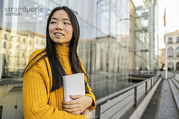Happy woman standing with book near glass building