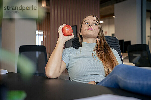 Bored businesswoman holding apple and sitting on chair in office