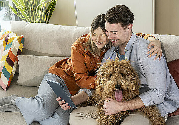 Glückliches junges Paar benutzt Tablet-PC und sitzt mit Hund auf dem Sofa im Wohnzimmer