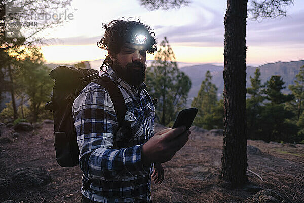 Mann trägt Stirnlampe und benutzt Smartphone bei Sonnenuntergang