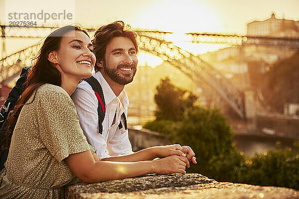 Lächelndes Paar hält Händchen und steht bei Sonnenuntergang vor der Dom-Luis-Brücke  Porto  Portugal