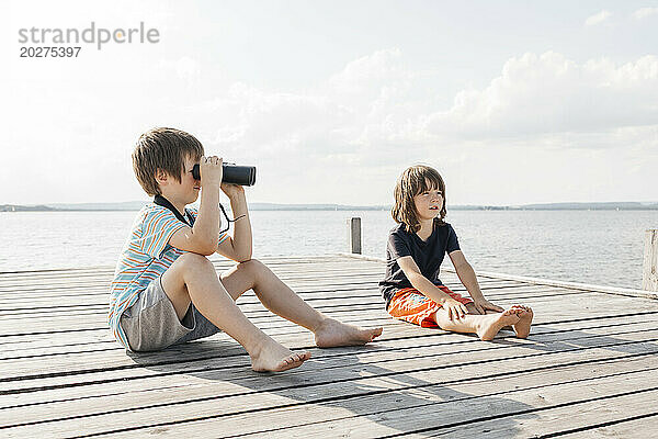 Junge schaut durch ein Fernglas und sitzt mit seinem Bruder am Pier am See