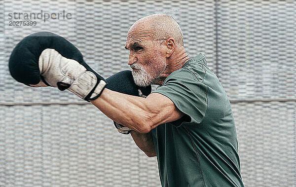 Selbstbewusster  aktiver älterer Mann übt mit Boxhandschuhen vor dem Zaun