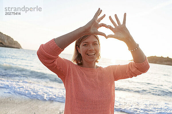 Glückliche Frau gestikuliert Herzform am Strand bei Sonnenuntergang