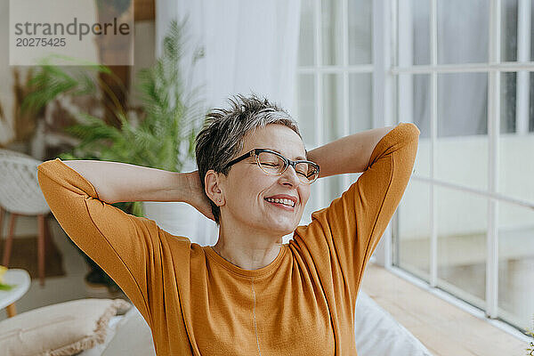 Happy woman with eyes closed and hands behind head at home