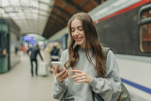 Lächelnde junge Frau benutzt Smartphone am Bahnhof