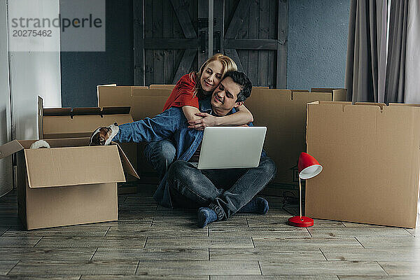 Glückliche Frau umarmt Mann  der mit Laptop neben Kartons sitzt
