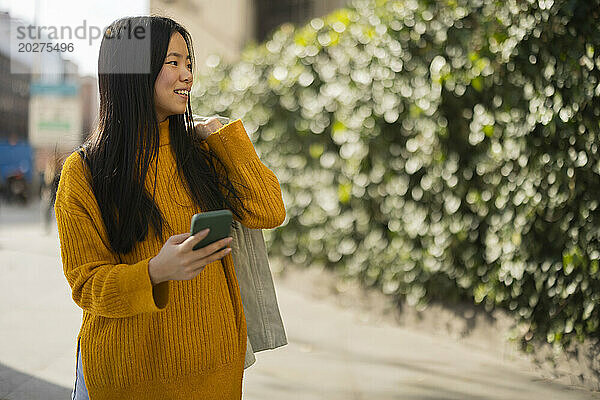 Glückliche junge Frau mit Tasche und Smartphone an einem sonnigen Tag