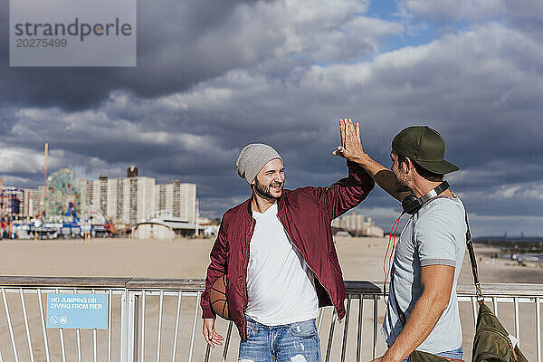 Glückliche Freunde geben High-Five beim Spaziergang in der Nähe des Geländers am Strand