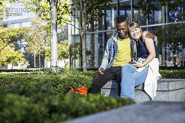 Lächelnder Mann sitzt mit dem Arm um einen Freund auf einer Bank im Park