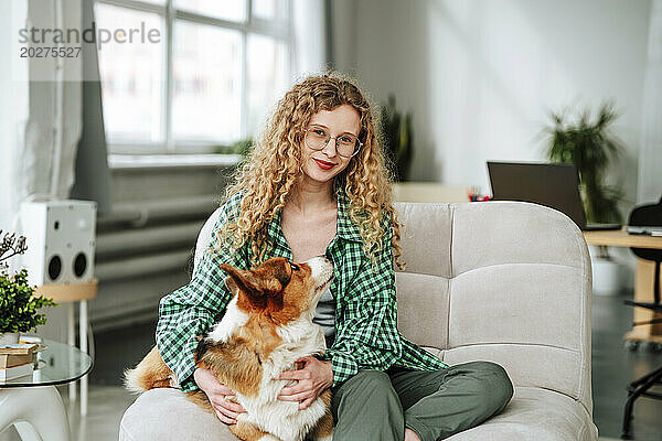 Lächelnde Frau sitzt mit Corgi-Hund auf der Couch zu Hause