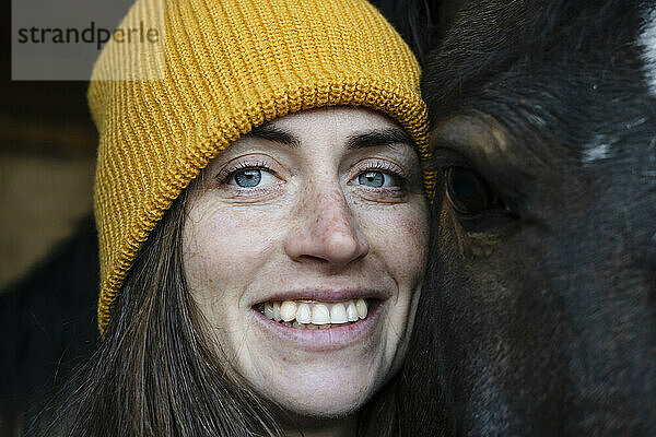 Lächelnde Frau mit Strickmütze neben geschecktem Pferd im Stall