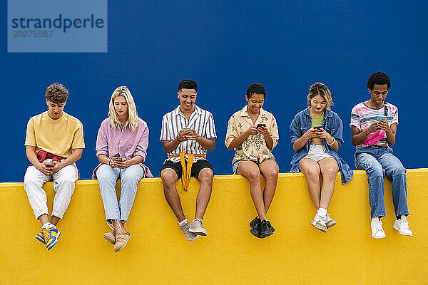 Fröhliche Freunde in bunter Kleidung sitzen auf Gelb und nutzen ihre Smartphones