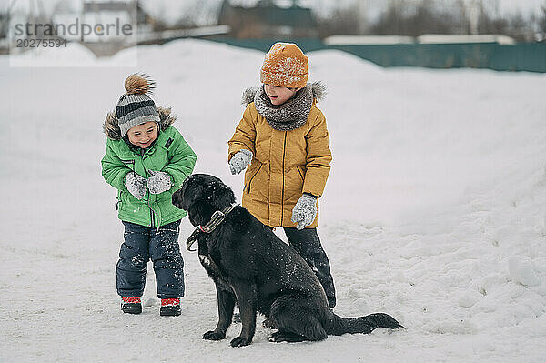Glückliche Kinder tragen warme Kleidung und spielen mit Hund im Schnee