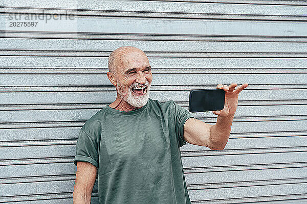 Ein pensionierter älterer Mann lacht über einen Videoanruf über sein Smartphone vor einem grauen Verschluss