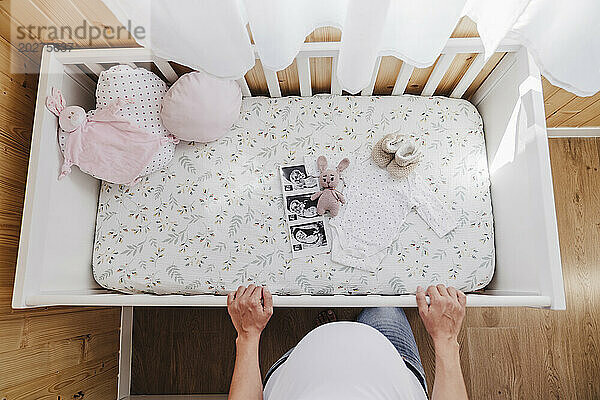Schwangere Frau steht mit Baby-Ultraschallfotos und Kleidung neben dem Kinderbett zu Hause
