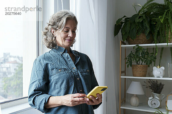 Reife Frau benutzt Smartphone am Fenster zu Hause