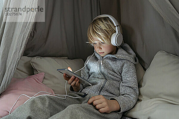 Junge trägt Kopfhörer und benutzt Smartphone im Baldachin zu Hause