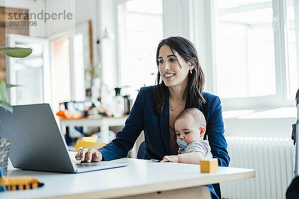 Lächelnde Geschäftsfrau sitzt mit ihrem kleinen Mädchen zu Hause und benutzt Laptop