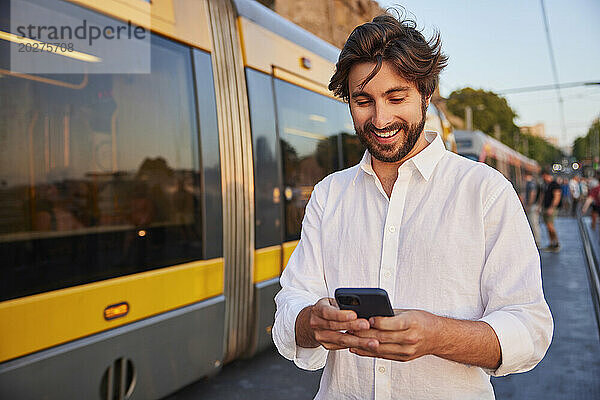Glücklicher junger Mann benutzt Smartphone in der Nähe der Straßenbahn