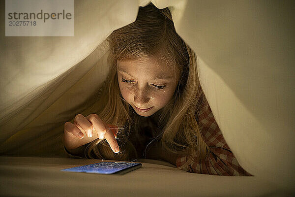 Girl using smart phone under blanket on bed at home