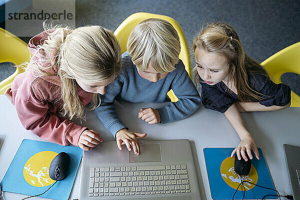 Blond boy sharing laptop with girls learning coding at desk in classroom