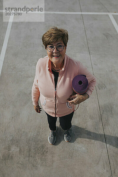 Lächelnde ältere Frau steht mit Trainingsmatte auf dem Parkplatz