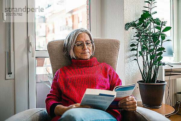 Reife Frau sitzt zu Hause im Sessel und liest ein Buch