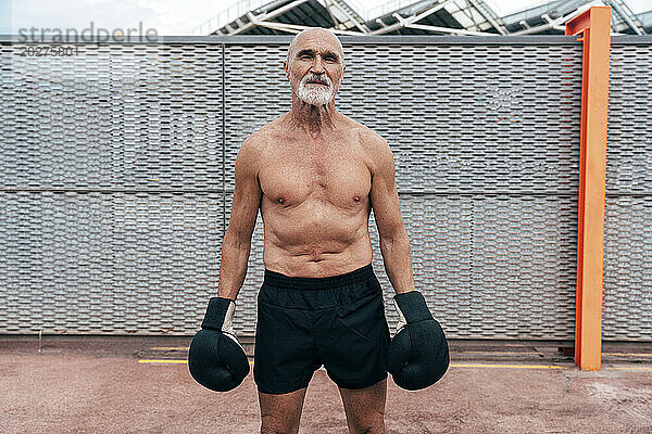 Oberkörperloser älterer Mann steht mit Boxhandschuhen vor dem Zaun